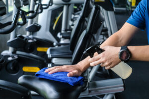 Cleaning professional wiping down gym equipment in a bright, modern fitness center with cardio machines and free weights visible in the background.